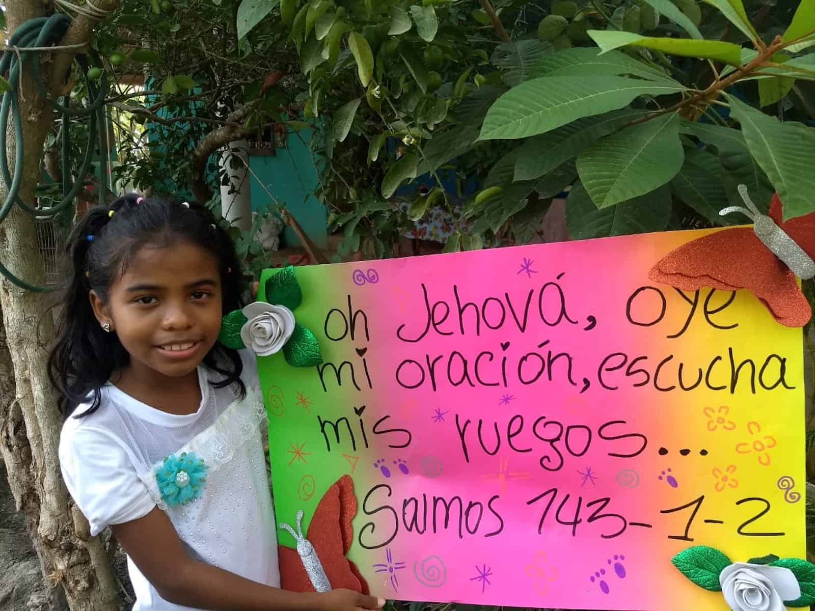 A girl holds a poster with a Bible verse written on it in Spanish.