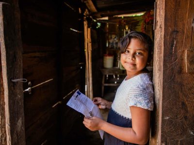 Young girl wearing a dress with a white bodice and a black skirt. She is standing in an open doorway of her home and she is holding a letter from her sponsor.
