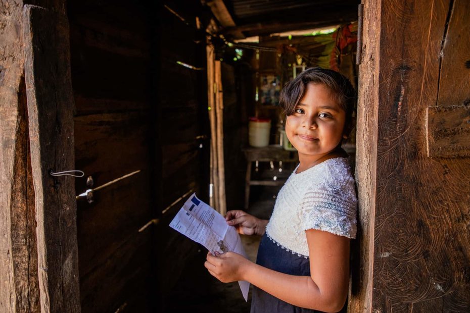 Young girl wearing a dress with a white bodice and a black skirt. She is standing in an open doorway of her home and she is holding a letter from her sponsor.