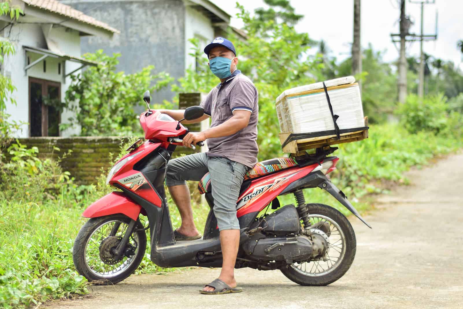 Julius is wearing a gray shirt and green pants, along with a blue face mask. He is sitting on a motorbike that he drives around his community to sell fish. His fish cooler is on the back of his bike.