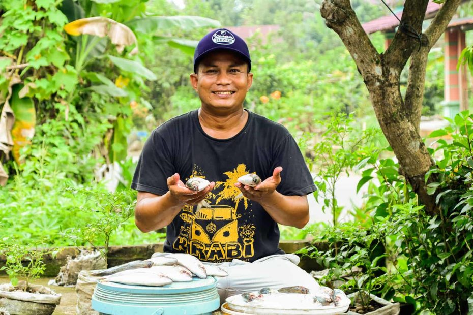 Julius is wearing a black shirt and a navy hat. He is holding two fish. In front of him are fish on a bucket that he will sell in his neighborhood. Behind him are trees.