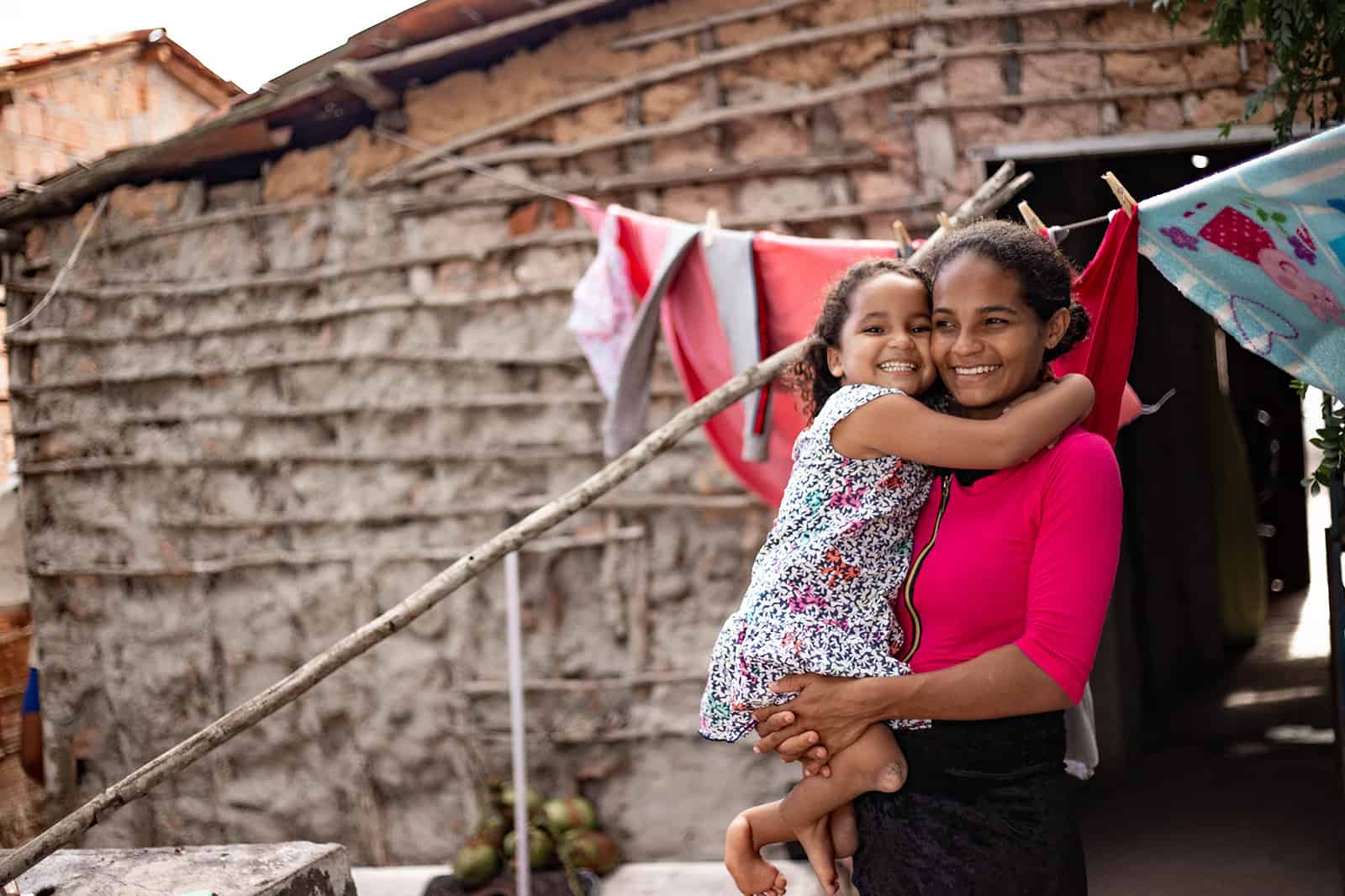 Maisa, in a blue and white dress, and her mother, Ana, in a hot pink shirt, are outside their home. There is laundry handing on a clothesline behind them. Ana is holding her daughter.