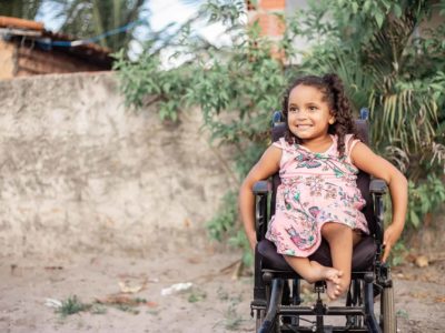 Maisa, in a pink dress, is sitting in her wheelchair on the road outside her house.