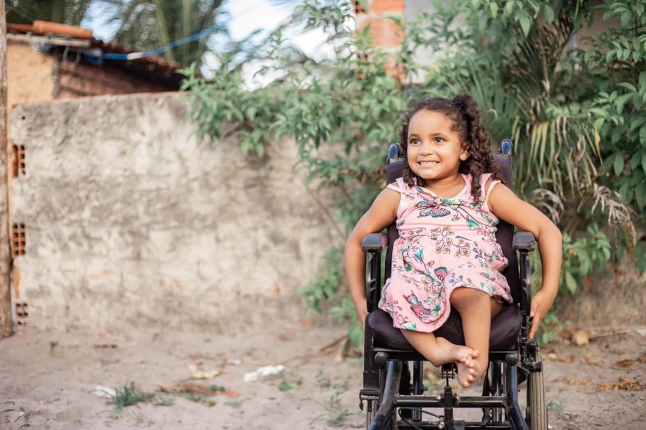 Maisa, in a pink dress, is sitting in her wheelchair on the road outside her house.