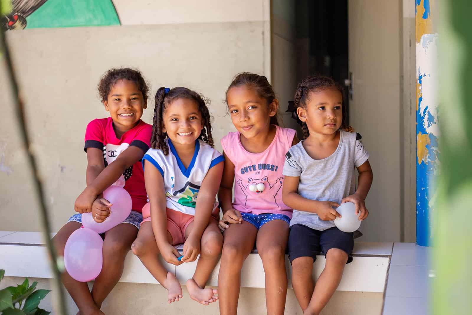 Maisa, in a blue and white shirt and pink shorts, is at the project sitting on a ledge with friends. One of the girls is holding balloons.