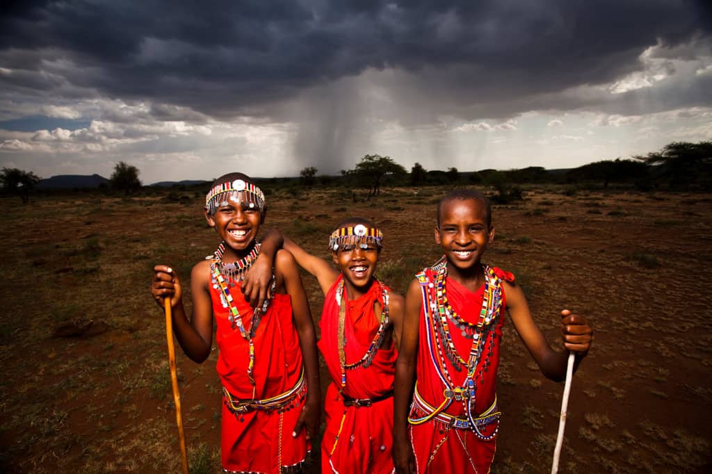 Masai in Najile, Kenya. A group of smiling boys in similar red clothes and headbands stand in a line in front of a dark sky and storm cloud.