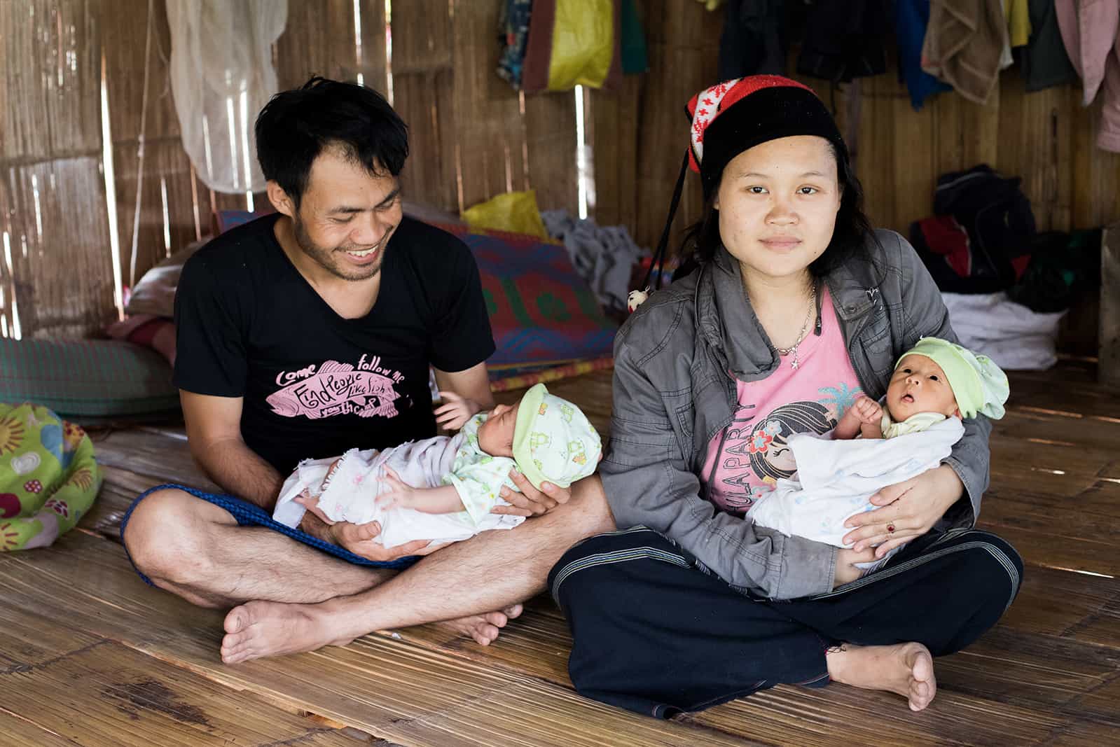 NoeDeMoo, Papala, and their twin baby girls, Phakaporn and Phakamon, sitting on the floor in their house. Papala is looking down at one of the twins smiling, and NoeDeMoo is looking at the camera.