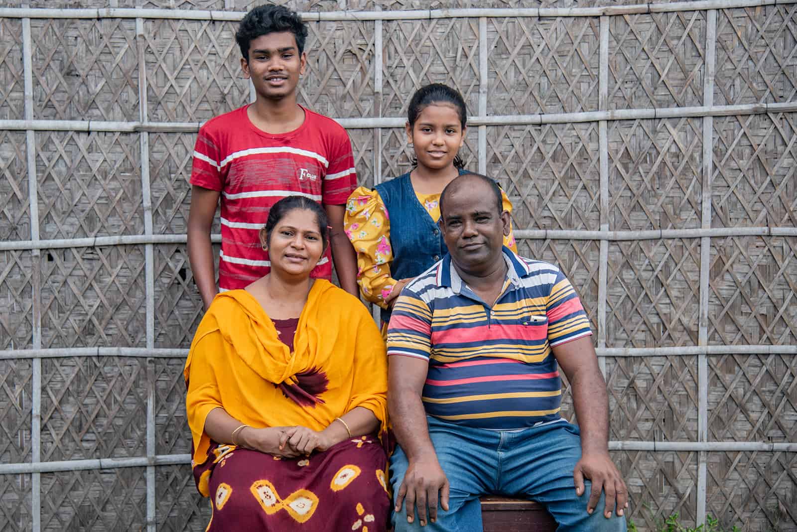 Sanjoy, in a red and white shirt, is with his family posing for a photo. His mother, in yellow, is sitting next to his father, Subhas. Sanjoy's sister is standing next to him. They are in front of a bamboo wall.