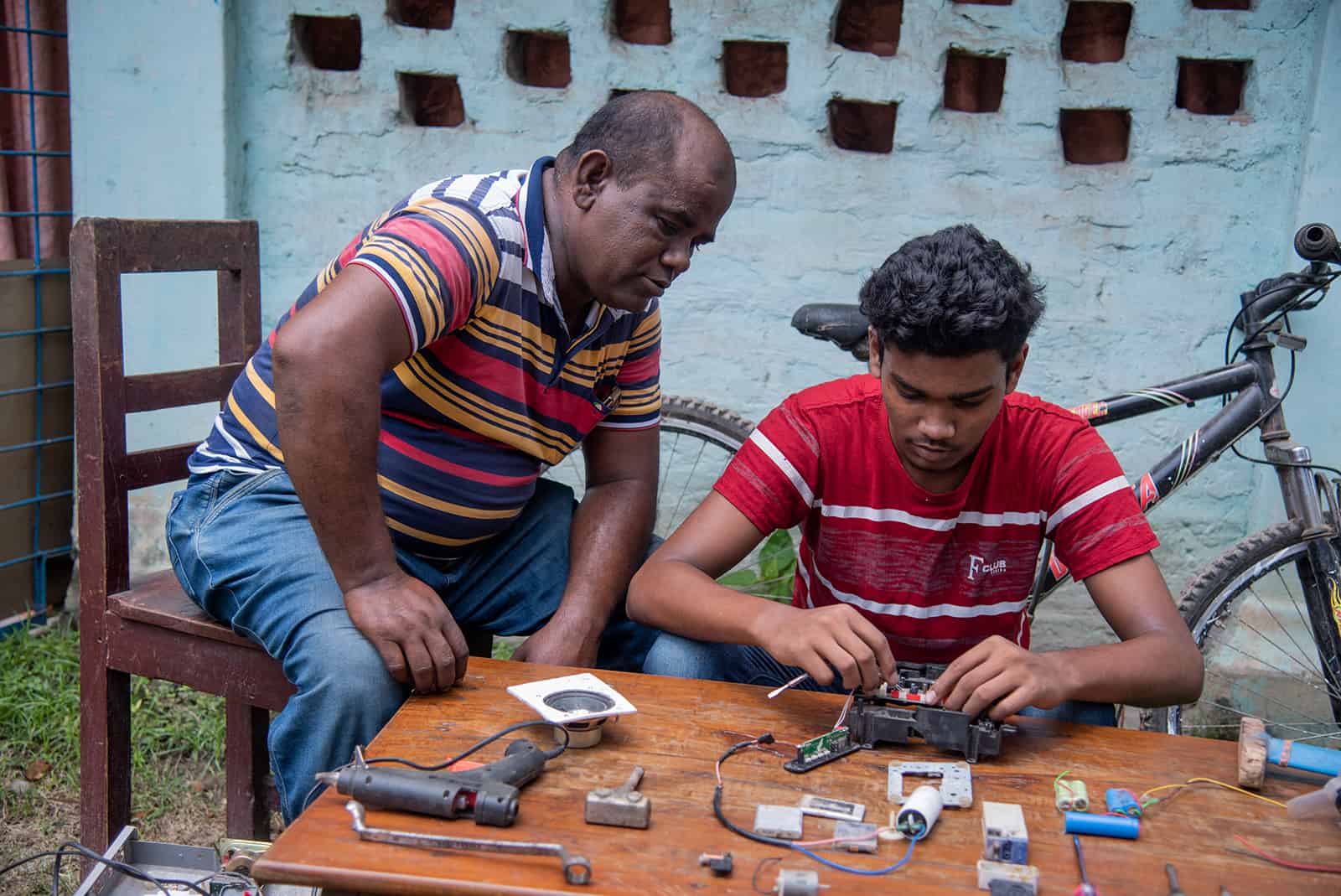 Sanjoy, in a red shirt, and his father, Subhas, in a red, yellow, blue and white shirt, are sitting at a wood table where Sanjoy is working on making a radio from a motherboard. There is a bike in the background.