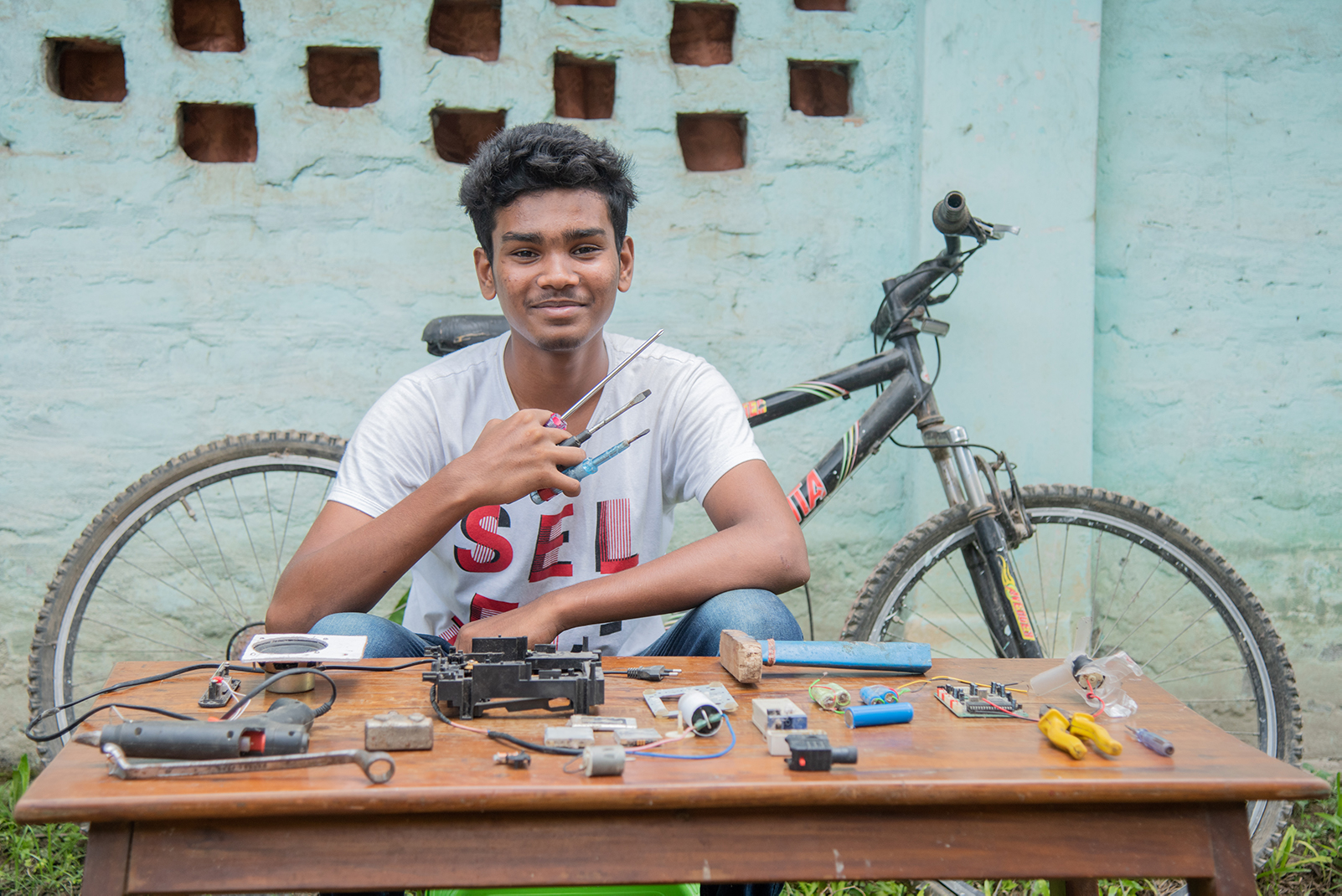 A young man sits at a table covered in tools