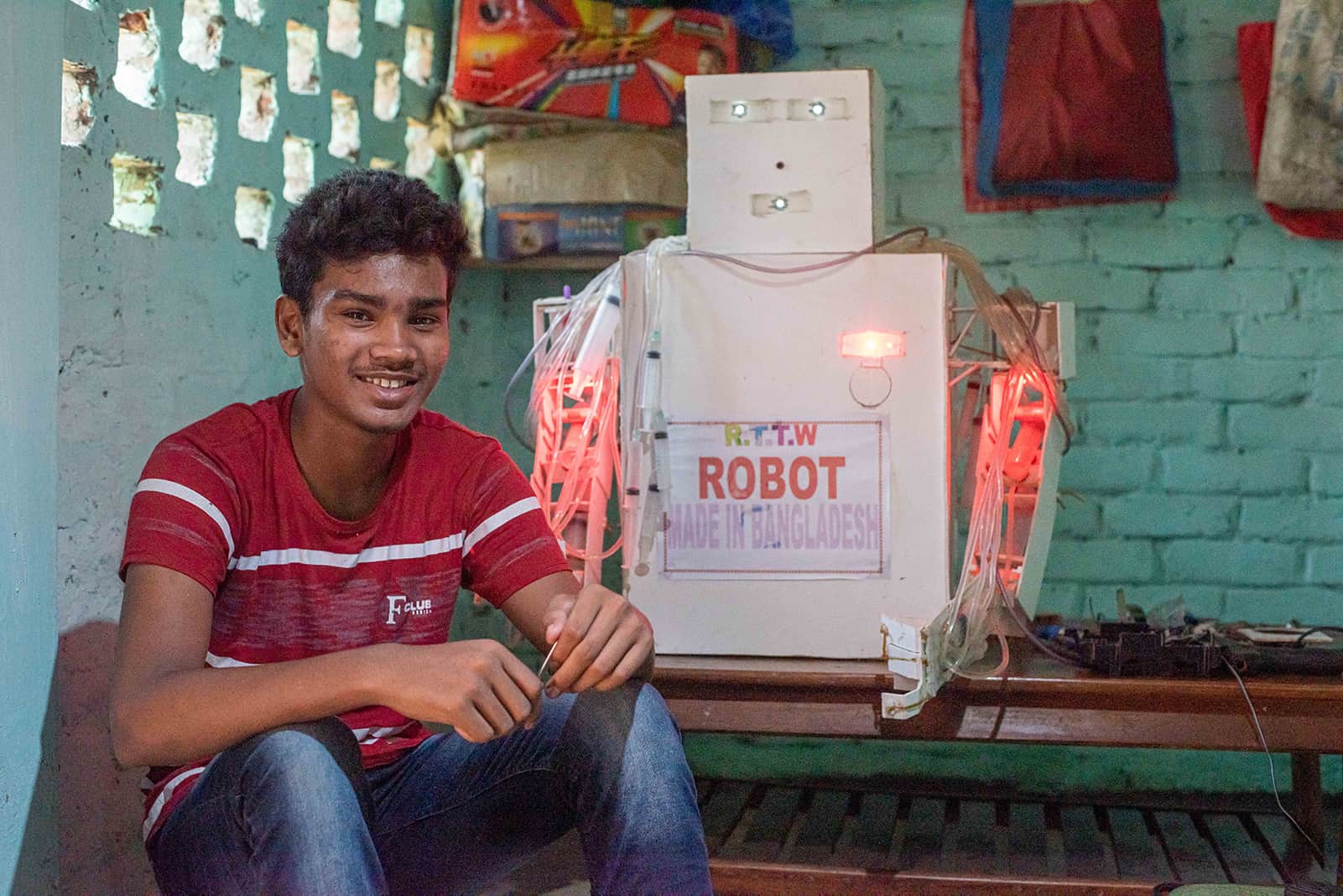 Sanjoy, in a red shirt, is sitting in front of the white cardboard robot he made for the science fair, where he came in second place.