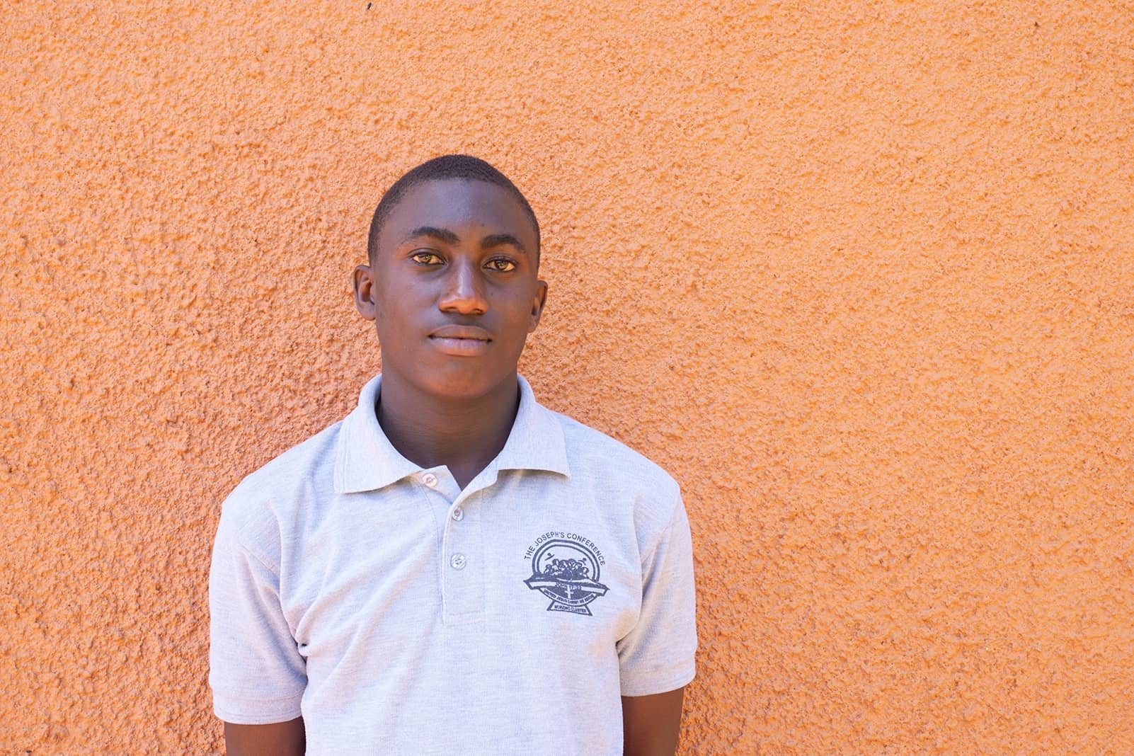 Young man wearing a gray shirt and is standing in front of an orange stucco wall.