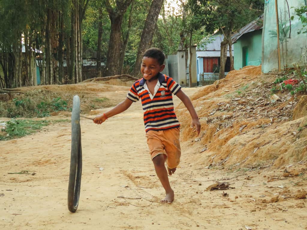 Boy wearing orange shorts with a red, blue, and white striped shirt. He is running down a dirt road with a bicycle tire and a stick.