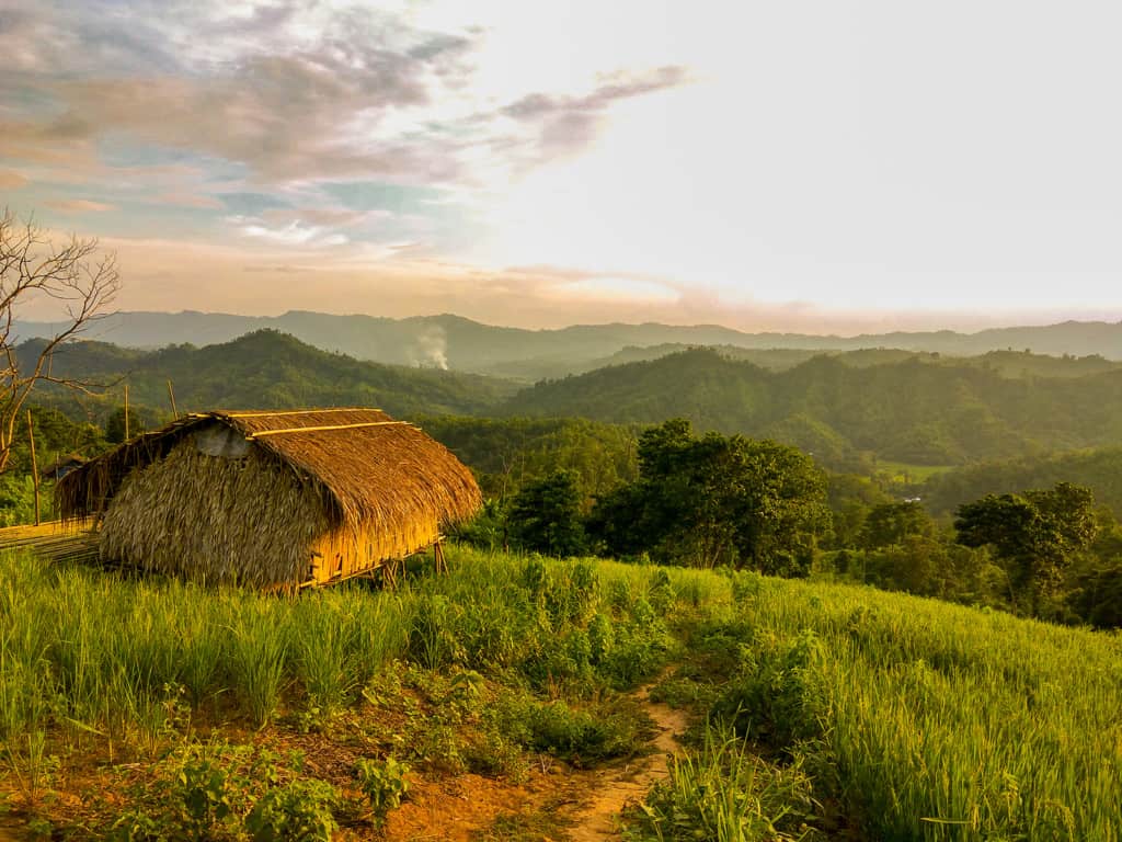 A light brown structure, building is in a field of tall grasses, crops overlooking green hills. There are clouds and smoke in the distance.