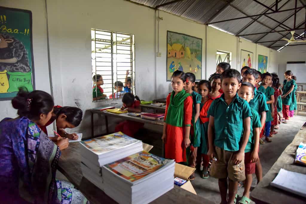 Children, most wearing red or green school dress or shirt uniforms, stand in two lines, facing towards a teacher's desk.