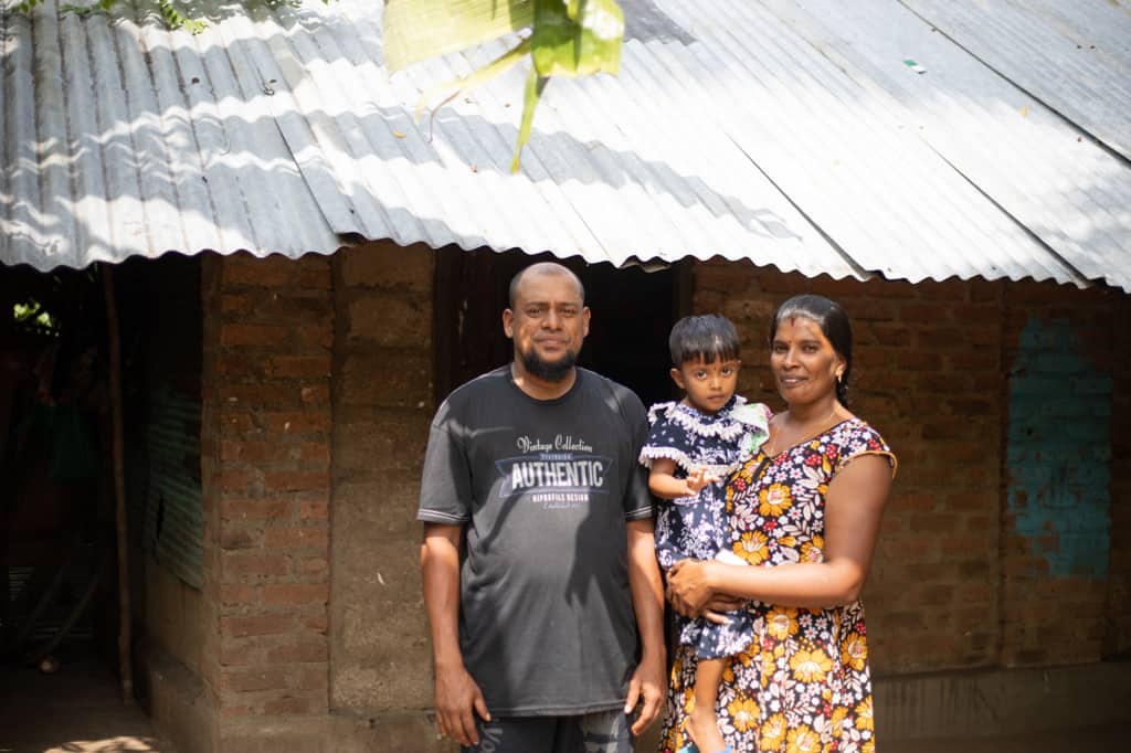 Densy is wearing a blue and white dress. She is being held by her mother, Jemalini, wearing a blue, white and yellow dress. Her father, Jenishtan, is standing beside them in front of their house which was submerged in three feet of water during the flood. 
