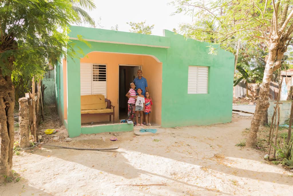 A family - 3 children and their father - standing on the porch of their mint green house.