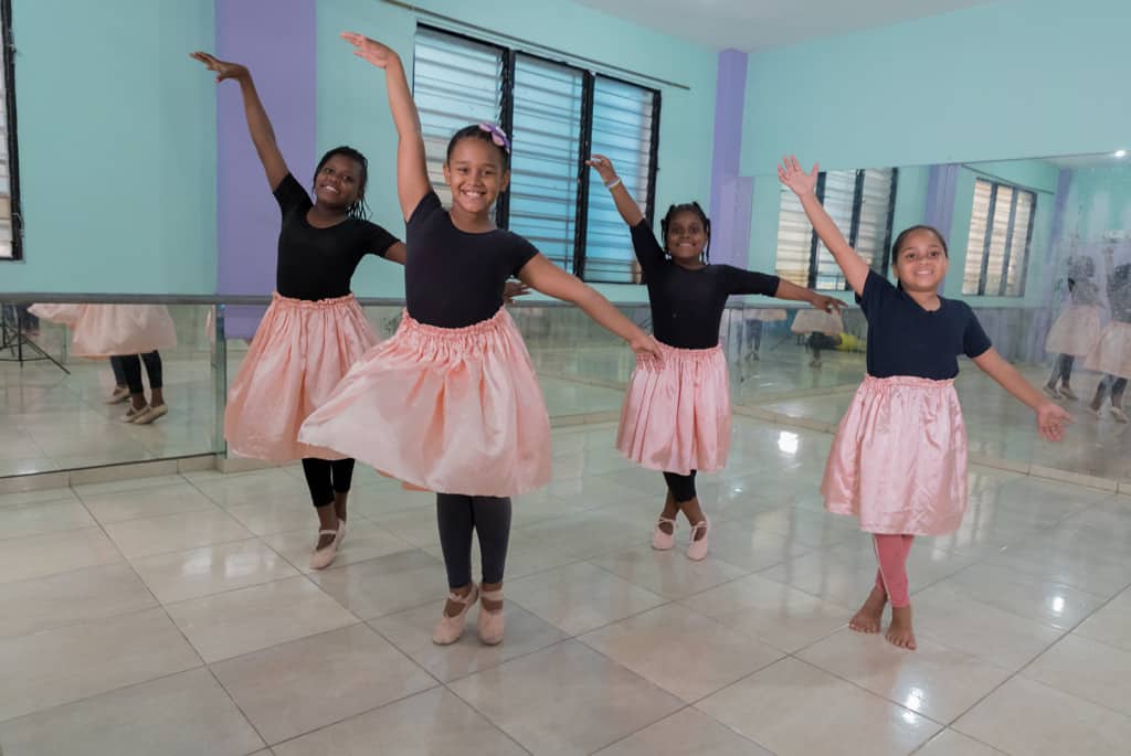 Four girls making eye contact and smiling while doing a dancing pose and smiling at the camera. They are all wearing black tee shirts, coral dancing skirts and light pink ballerina dancing shoes.