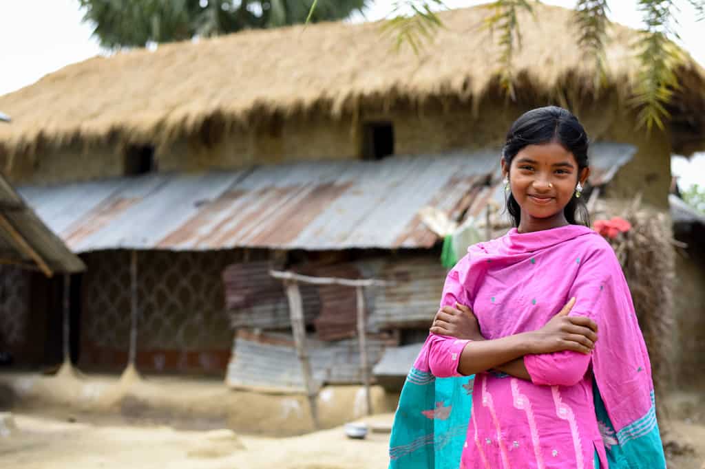 A girl wearing pink and blue clothing in front of her home made of corrugated metal, grass, hay and repurposed materials. Her arms are crossed.