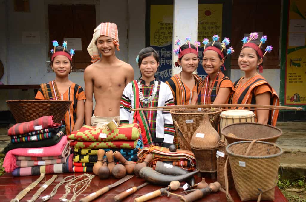 A group of children, boys and girls, teens, teenagers, youth, smile wearing beads, colorful clothes, hats, turbans, sarongs, orange and black cloth fabric dressed in costumes of region and ethnic background, stand together outside at a culture show day at project behind a table of items from their local region and country.