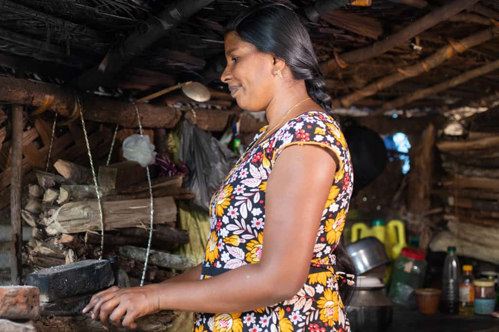 Jemalini is wearing a blue, white, and yellow floral dress. She is cooking in her kitchen on the top of a barrel. Behind her, there is firewood used for cooking tied up to the wall so it will not get wet.