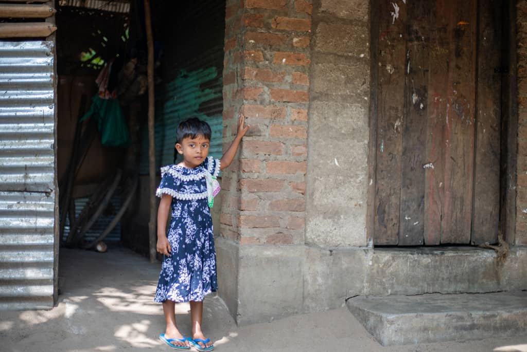 Densy is wearing a blue and white dress and is standing in front of her home. She is putting her hand up on the brick to show where the water was.