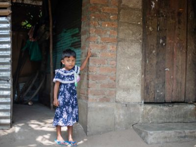 Densy is wearing a blue and white dress and is standing in front of her home. She is putting her hand up on the brick to show where the water was.