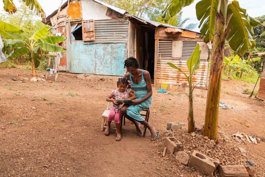 Young girl sitting in front of her house next to her mother who is reading the Bible to her.