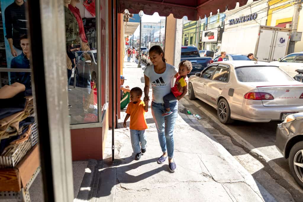 A boy, wearing an orange shirt, walks down the sidewalk with his mother, who is also carrying another child in her arms as they go shopping at the store and market.
