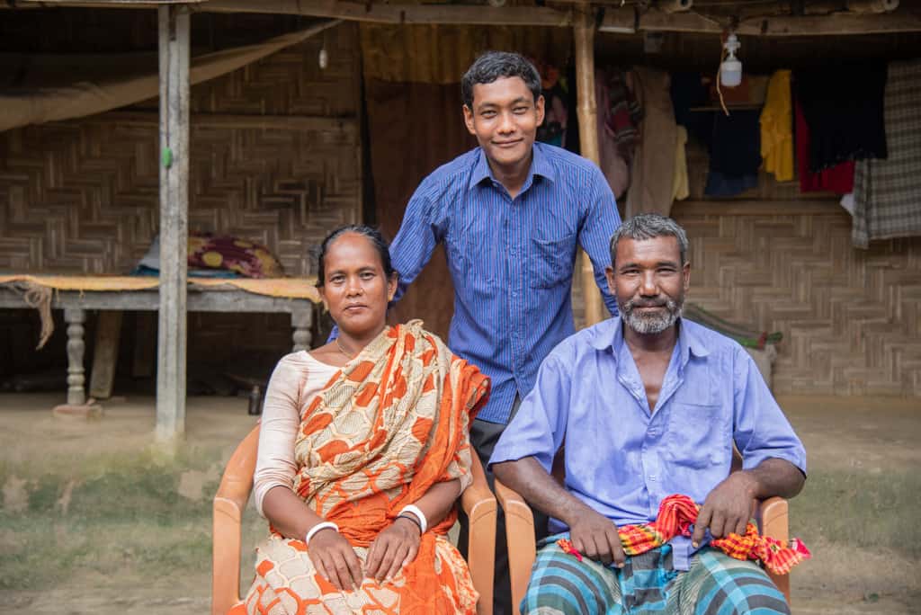 Lipon is wearing a blue shirt and gray pants. He is with his parents in front of their mud hut.