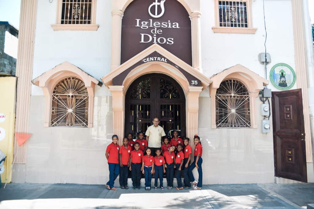 A pastor in a beige shirt standing in front of his church surrounded by children wearing red polo shirts.