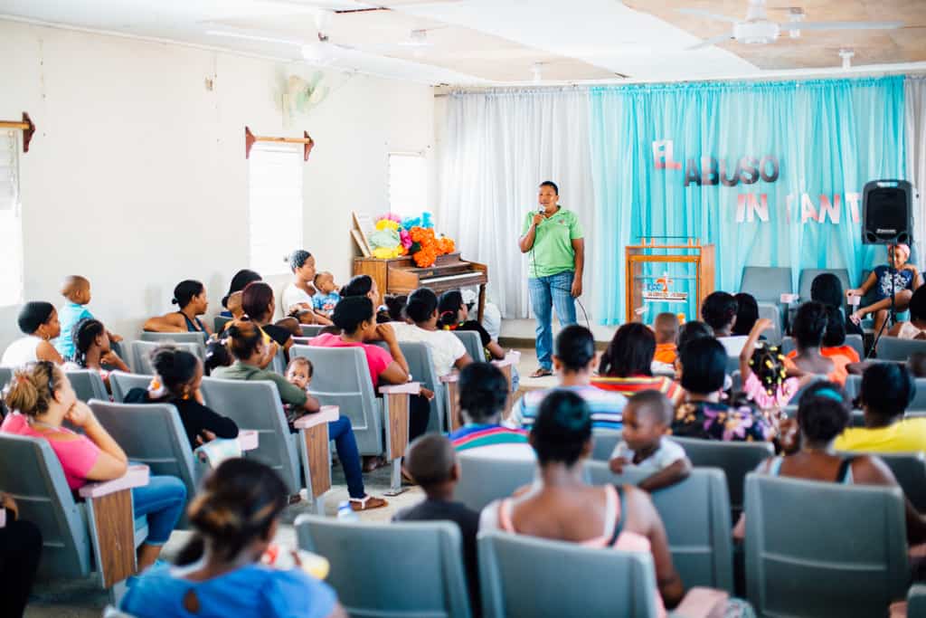 The backs of moms and children sitting in a room looking at a person in green shirt and blue jeans holding and speaking into a microphone on a stage. The walls are white and there is a blue and white curtain on the stage.