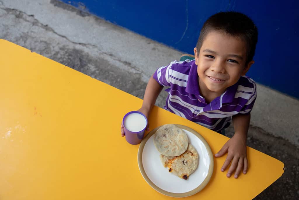 Smiling boy wearing a purple and white striped shirt sitting at a table with Pupusas and a cup of milk