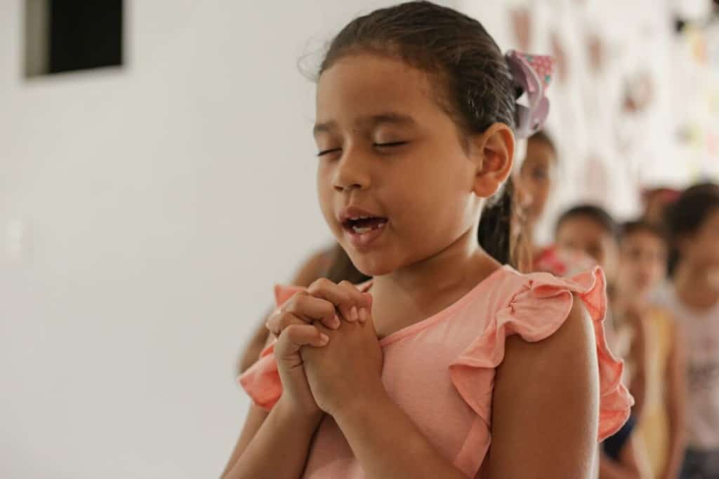 A Brazilian girl wearing pink prays with her eyes closed and hands clasped