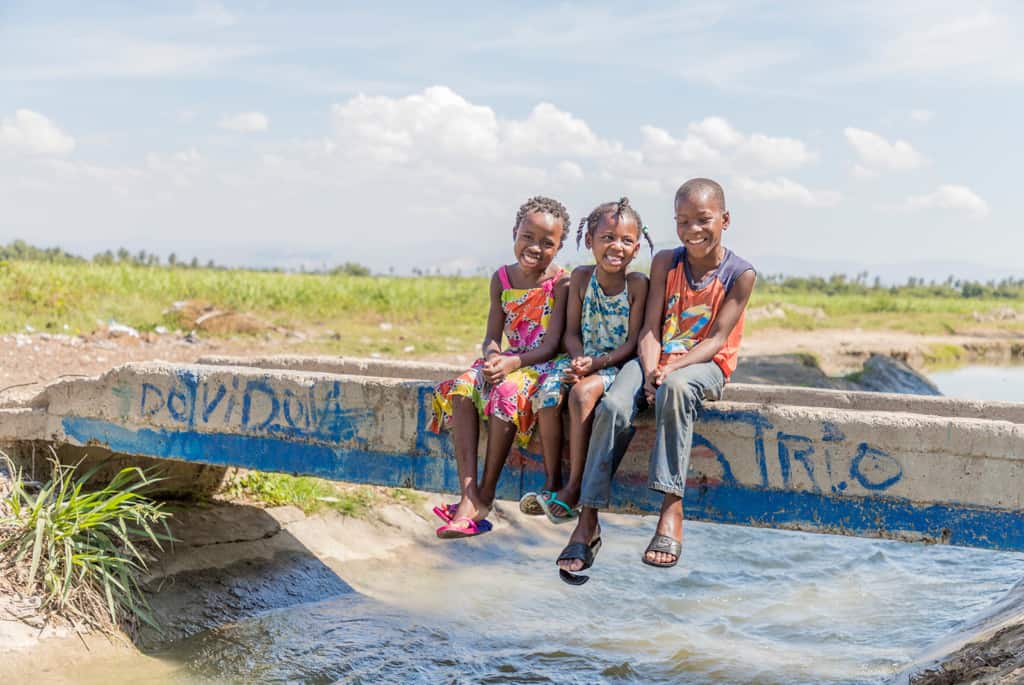 The girl on the left is wearing a yellow dress with blue flowers, the girl in the middle is wearing a white dress with bright colored flowers in green, yellow, orange, and pink, and boy on the right is wearing an orange sleeveless shirt and khaki pants. They are sitting on the bridge that connects the field with the community. 