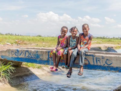 The girl on the left is wearing a yellow dress with blue flowers, the girl in the middle is wearing a white dress with bright colored flowers in green, yellow, orange, and pink, and boy on the right is wearing an orange sleeveless shirt and khaki pants. They are sitting on the bridge that connects the field with the community.