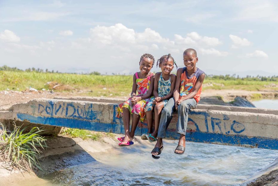 The girl on the left is wearing a yellow dress with blue flowers, the girl in the middle is wearing a white dress with bright colored flowers in green, yellow, orange, and pink, and boy on the right is wearing an orange sleeveless shirt and khaki pants. They are sitting on the bridge that connects the field with the community.