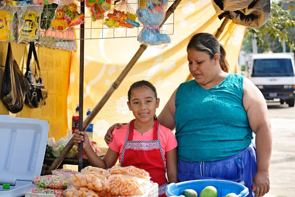A girl wearing a pink shirt and red small apron smiles standing besides her mother who is wearing a sleeveless blue tank shirt at their small food open market stall selling fruits, homemade food, and drinks.