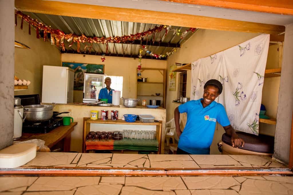 An adult woman wearing a blue shirt stands in a market shop store smiling looking out of the counter and there is an adult woman behind her looking out from an open window inside the shop.