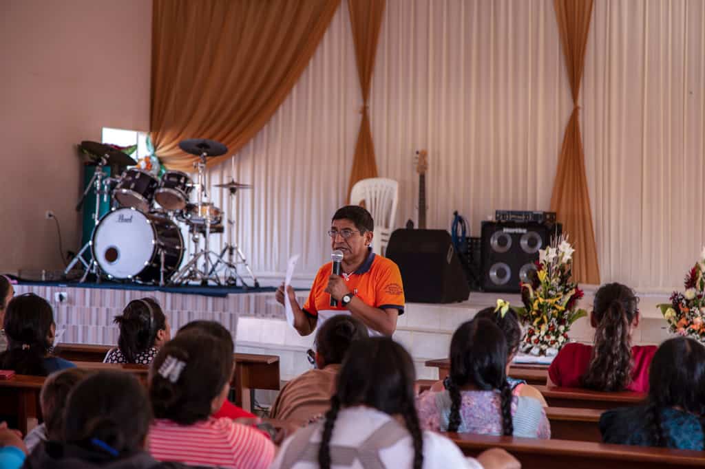 A man, in an orange shirt, holds a microphone and stands in front of a group of women sitting in pews. There is a stage with a drum set behind him.