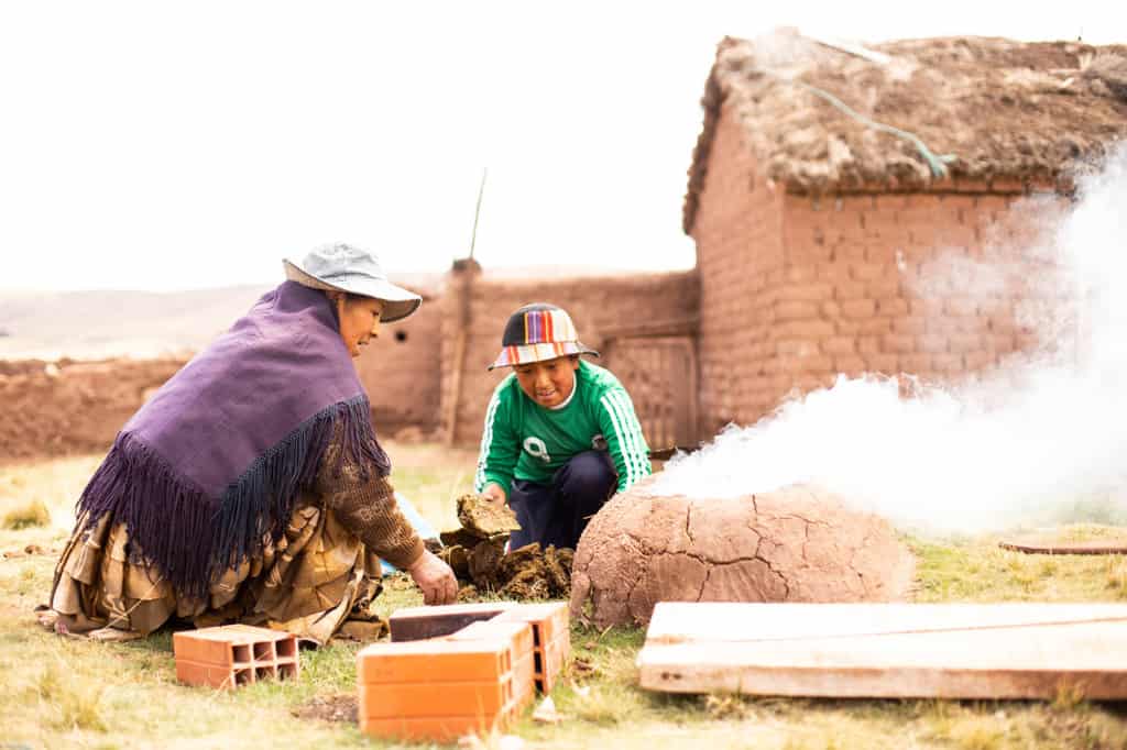 A boy and his grandmother are working outside. She is wearing a purple shawl and he is wearing a green long sleeved shirt and a colorful hat. There is a brick building in the background.