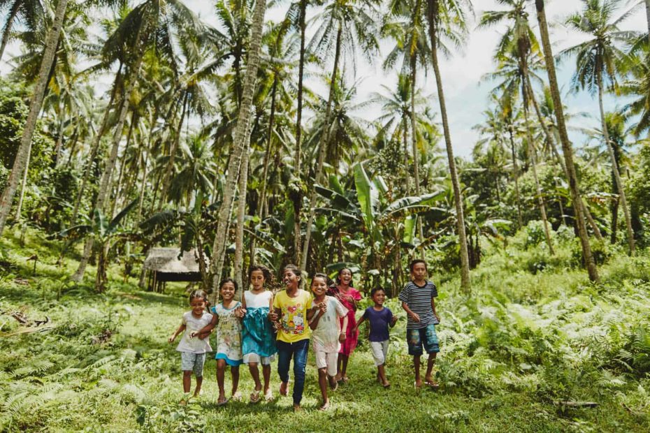 Happy, smiling, laughing children, boys and girls, males, females run outside in the grass surrounded by tropical, palm trees wearing blue, yellow shirts, white shorts, blue and white dress.