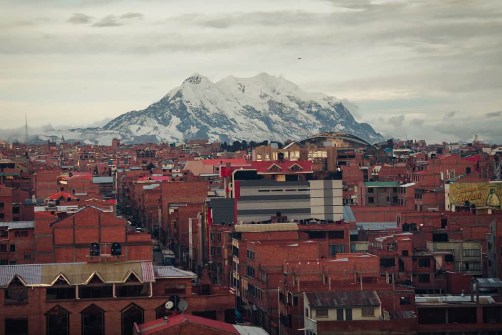 Brown brick buildings are in the foreground and a tall snowcapped mountains rises in the background  in the city of El Alto, La Paz.