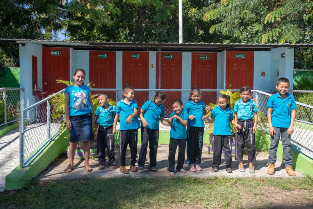 A group of beneficiaries, all wearing blue tee shirts, laughing and smiling all together. Behind them are the new restroom facilities with the first toilets in the community that flush with water.
