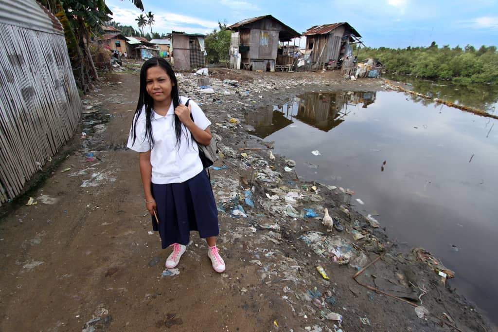 Girl wearing a white shirt, walks through her neighborhood and community, dirt road beside poor poverty homes, on her way to school. She is walking beside a water filled stagnant unhealthy unsanitary, pool of polluted water with garbage and trash strewn along the dirt road.