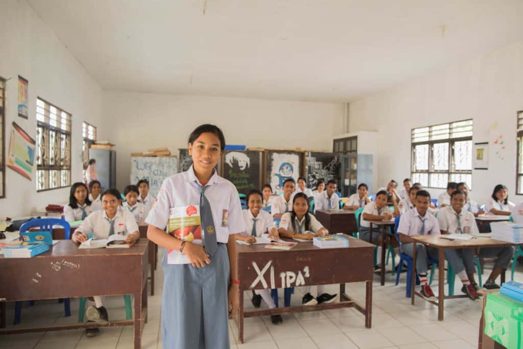 Girl wearing a gray skirt and white shirt. She is holding a book and is standing in front of her class at school. There are several students behind her all sitting at desks.