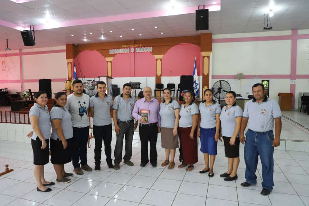 Pastor Pedro Segovia and the Church’s staff are in the sanctuary. Most of them wearing a grey polo shirt and Pastor Pedro is wearing a purple button-down shirt. The background is the altar where the Pastor preaches to the congregation and the worship band leads.