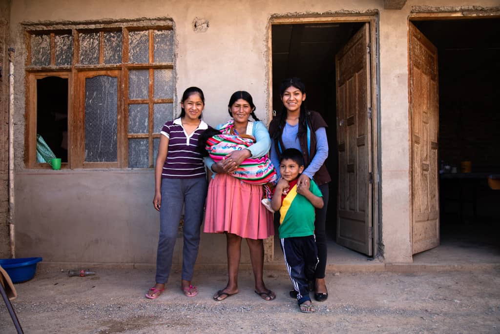 Jimena, her mother Esutaquia, her sister Tais, her 4-year-old brother Erwin, and the one-year old twins, are standing outside together. There is a tan building behind them.