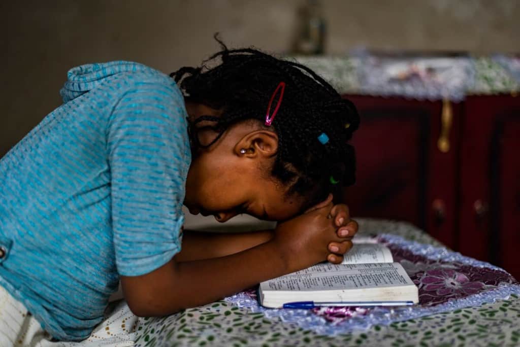 A young girl wearing a blue shirt prays over an open Bible