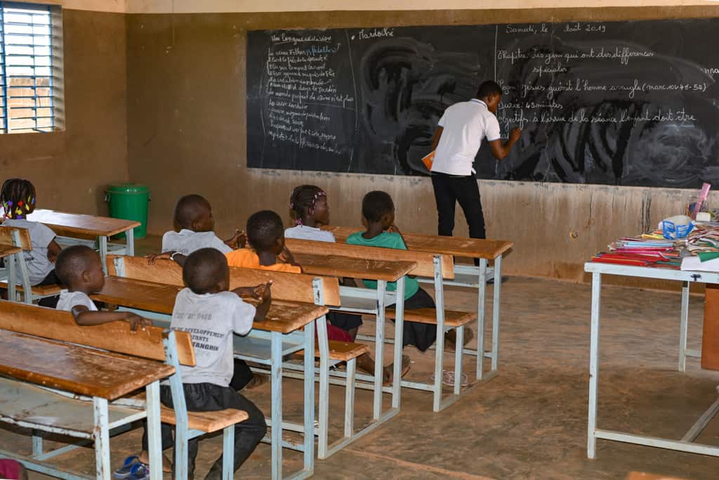 Man wearing a white polo shirt writing on a chalkboard with children at desks watching.